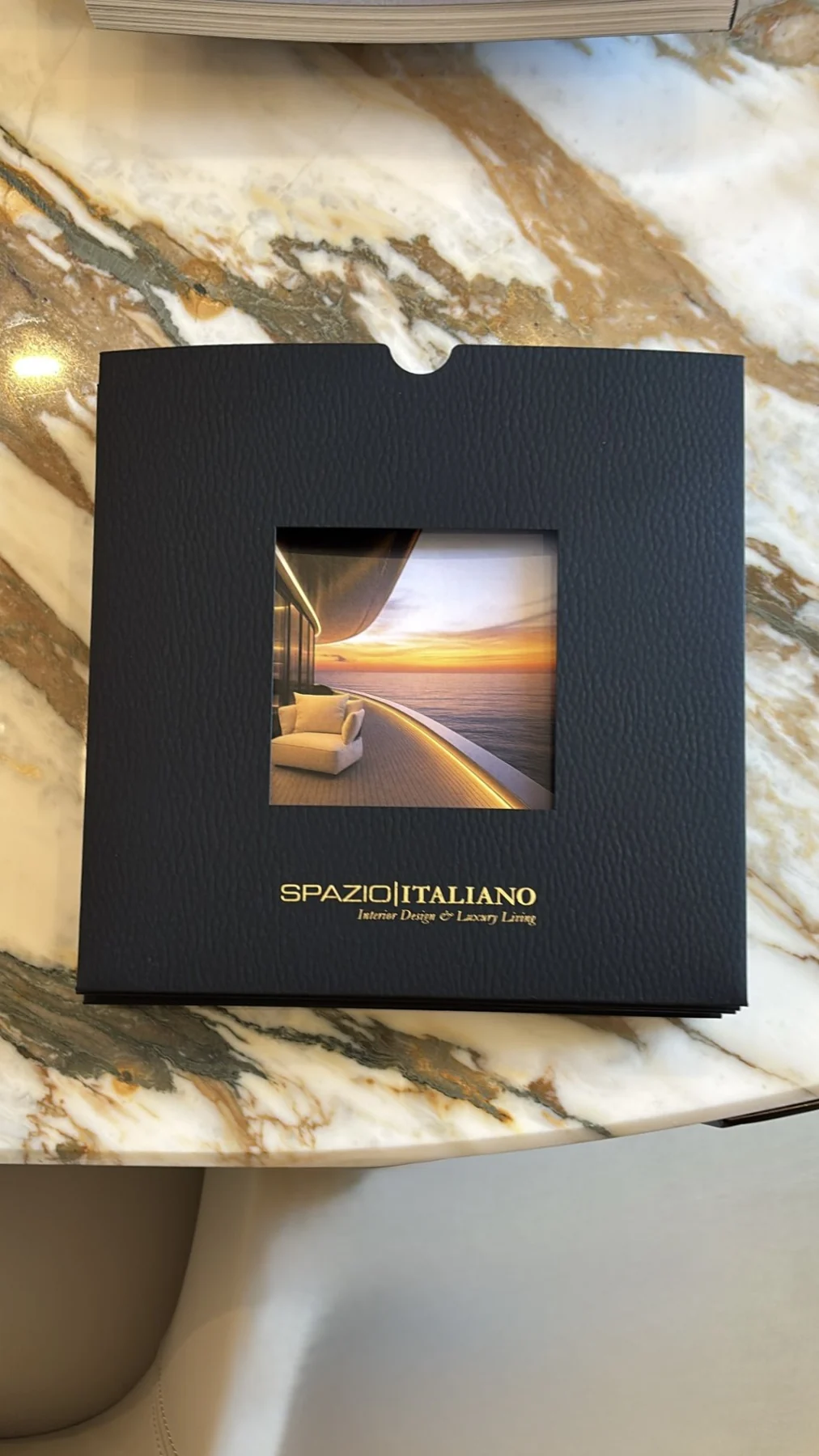 Brochure Spazio Italiano dans sa pochette noire à fenêtre, avec un visuel de yacht au coucher du soleil et une signature dorée.
